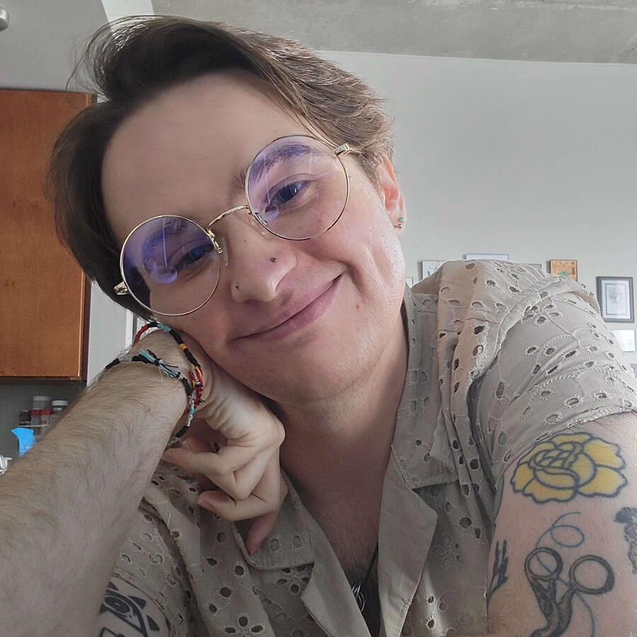 Orion A selfie of Orion, a white trans-masculine man with light brown hair, large round glasses, and two nostril piercings. He wears a beige lace short sleeved button down shirt and several bracelets. He is smiling and resting his cheek on his hand.
