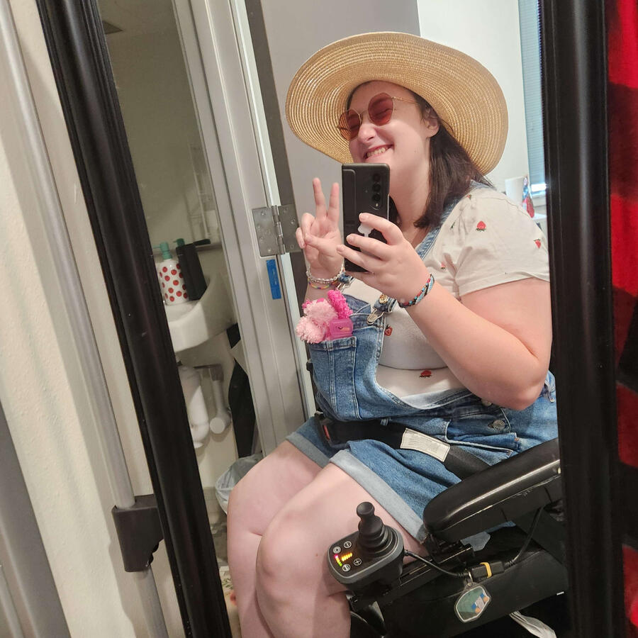 Sadie A mirror selfie of Blake, a white femme. It is wearing overalls, a white t-shirt with strawberries, and a straw hat. They are sitting in their wheelchair, making a peace sign, and smiling. A pink dog beanie baby is in the pocket of its overalls.