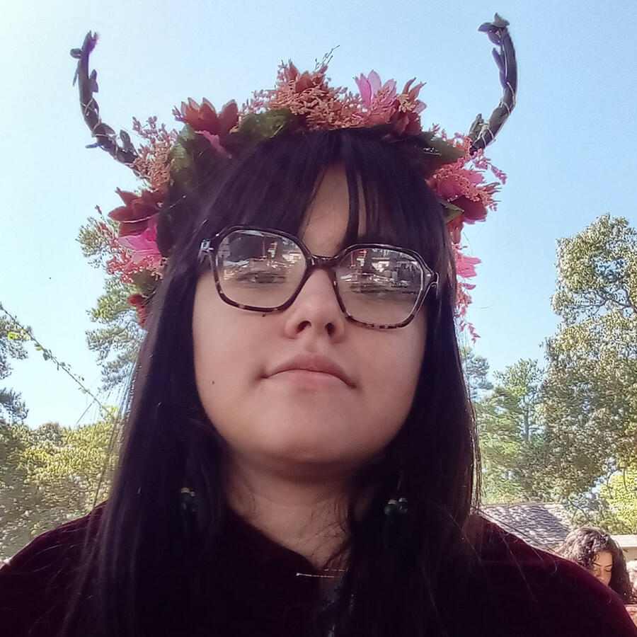 An outdoor selfie of Kamila at the Texas Renaissance Faire. She is wearing a headband with pink and dark red flowers with antlers as well as a dark red cape and circular black glasses.